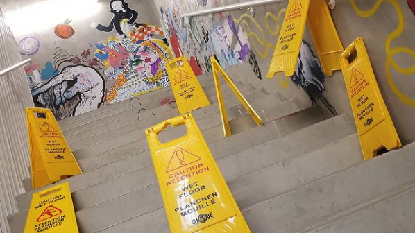 Seven yellow plastic “wet floor” hazard signs are placed haphazardly, blocking a set of stairs in a concrete hallway. The walls are painted with colourful murals.