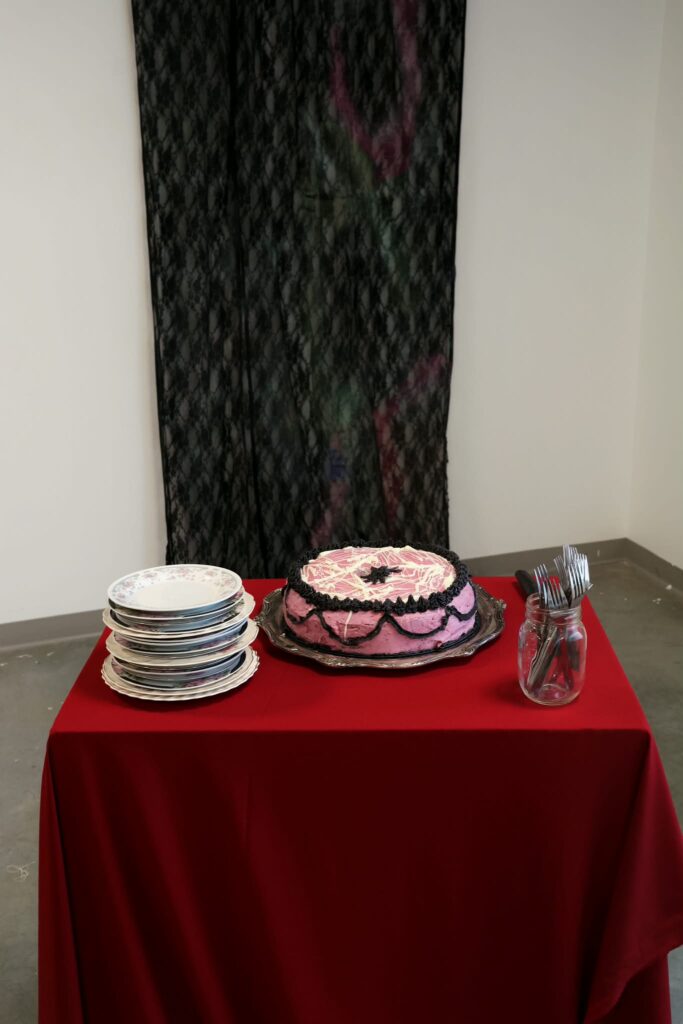 A triple layer gluten free vanilla cake with fresh raspberries running through it, raspberry coulis, white chocolate whipped cream, white chocolate chips, blueberry and raspberry buttercream with melted white chocolate and charcoal buttercream decoration on silver serving tray, knife, cup of forks, serving utensil, pink floral-patterned plates, displayed on plinth covered with red fabric.
Cut and served during presentation.