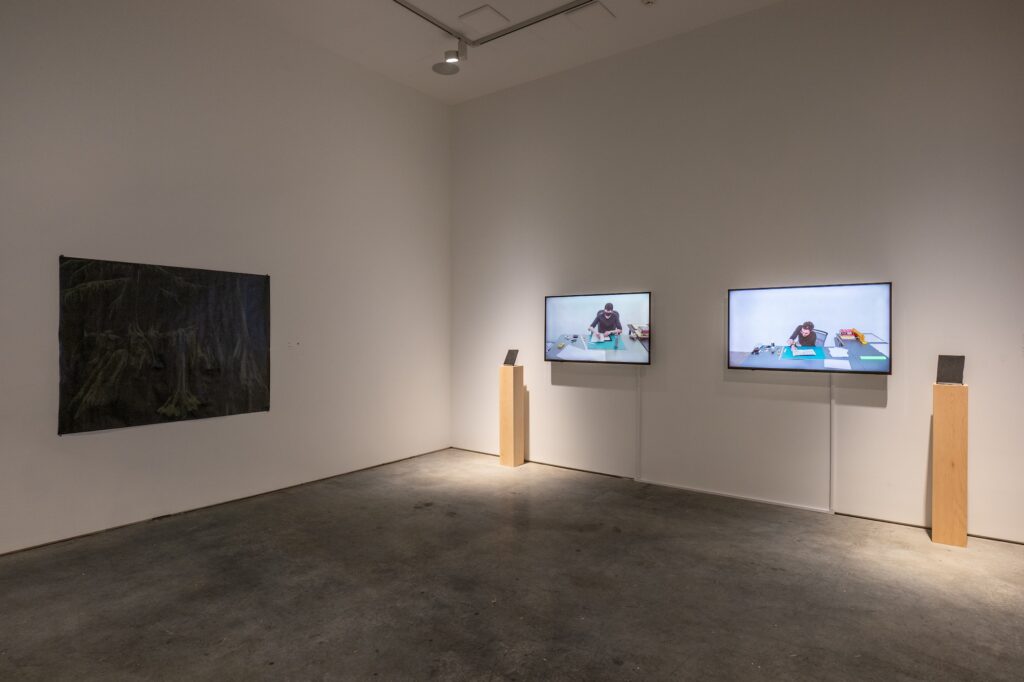A dark gallery room with two screens mounted on the wall, and plinths besides them displaying small two-dimensional items. A separate dark rectangular form hangs on the adjacent wall. The works and the lighting create a focused, intimate environment.
