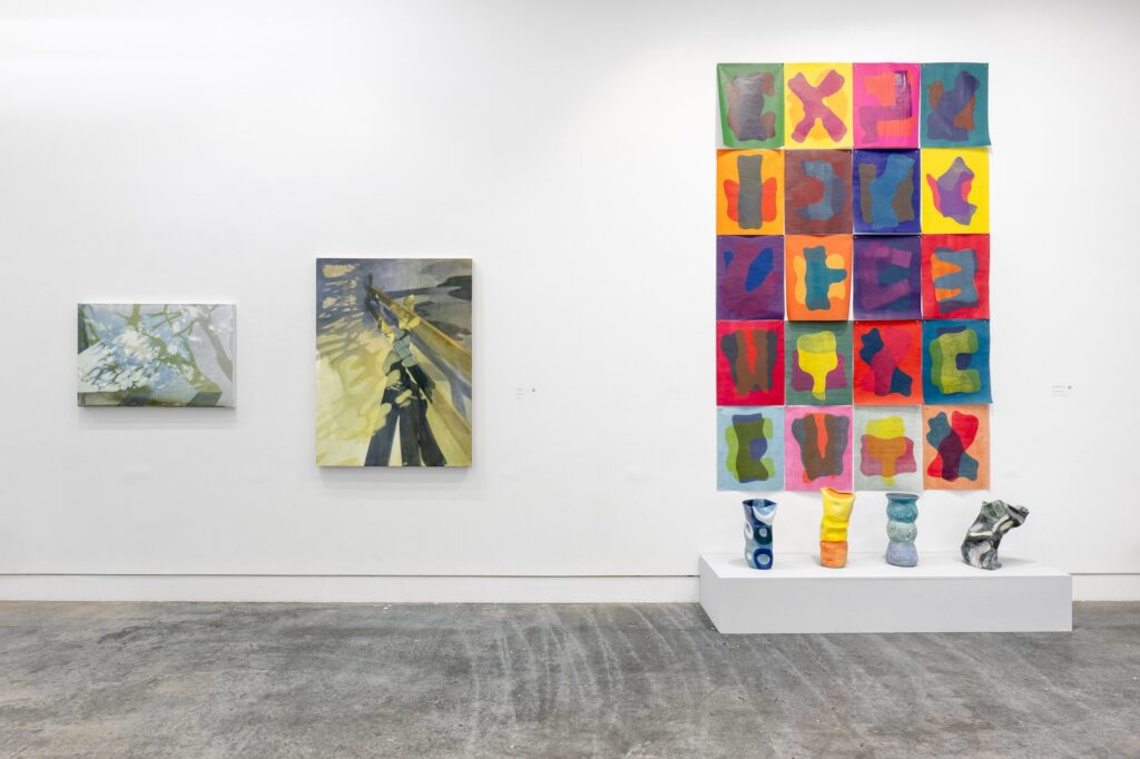 Shown close up, multiple large two-dimensional works are displayed on the gallery wall. A collection of sculptures stands on a low plinth on the floor beneath one of the paintings, echoing its bright colour scheme. 