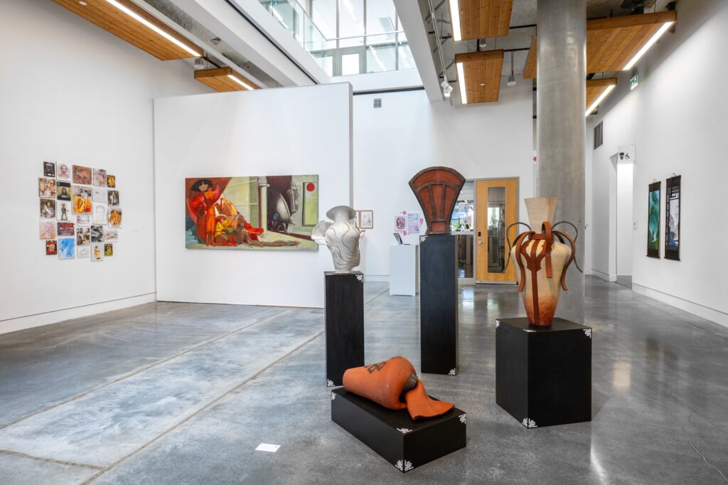 A series of distinguished, large vessels stand atop black, detailed plinths. Multiple two-dimensional works are displayed on the gallery walls, featuring images of different sizes, colours, and mediums.