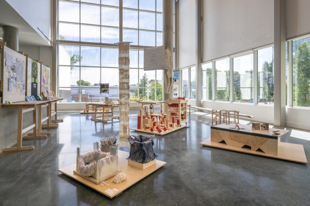 A bright gallery room with window walls and high ceilings displays an array of design projects. The works are displayed on tables, platforms, and some hang from the ceiling. The room is filled with works ranging from furniture, textile objects, paper booklets, and posters. Most of the works are wooden, or share a similar warm, natural tone.