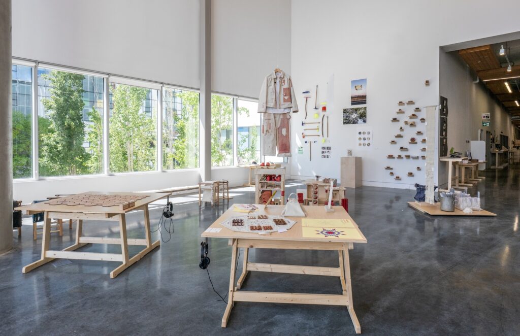 A bright gallery room with window walls and high ceilings displays an array of design projects. The works are displayed on tables, platforms, and some hang from the ceiling. The room is filled with works ranging from furniture, textile objects, paper booklets, and posters. Most of the works are wooden, or share a similar warm, natural tone.