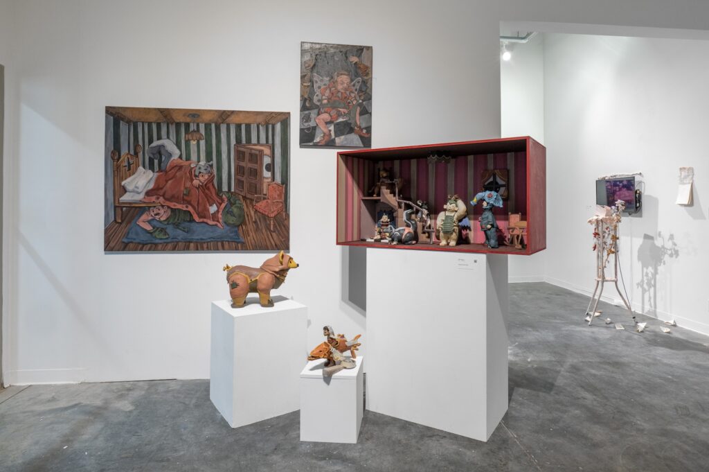 Gallery wall displays a cluster of sculptures and paintings, creating a world-like feeling of characters and figures. The sculptures of different creatures stand atop plinths, with some of them inside a horizontal striped box. The paintings on the wall echo the dark, subdued, and earthy tones of the sculptures. Other sculptures stand farther in the background.