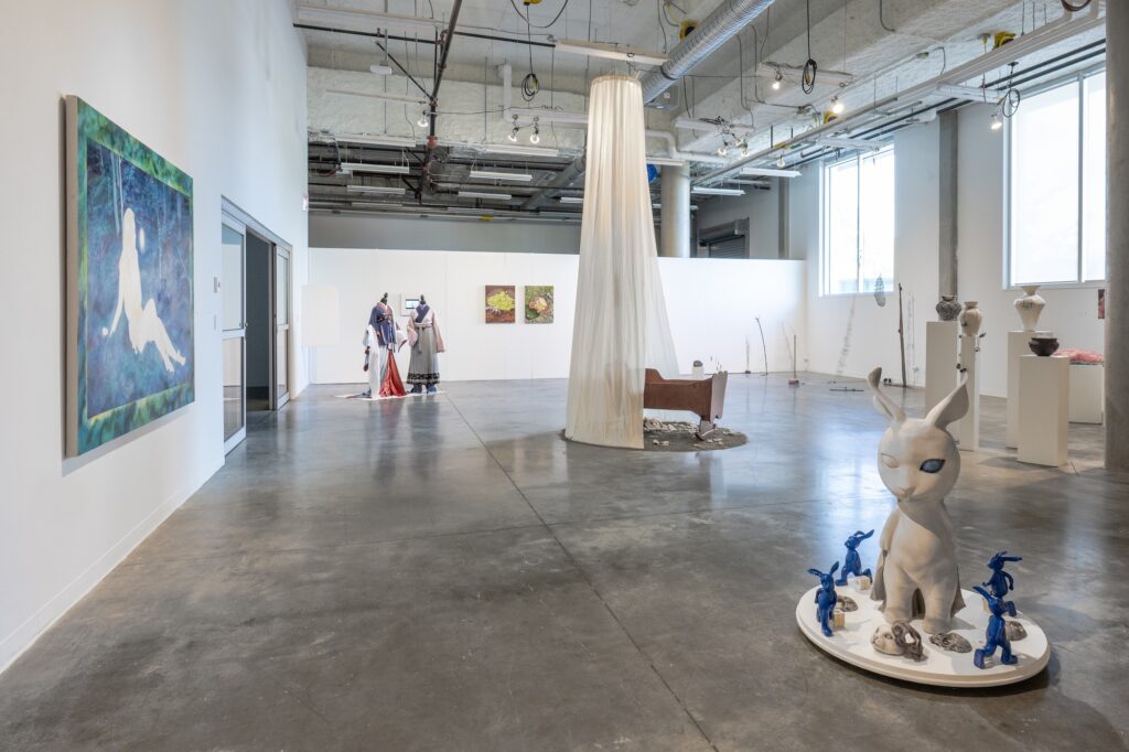 A bright gallery room shows an array of sculptures and paintings, ranging in scale, medium, and themes. In the centre of the room stands the tallest sculpture, which has a long flowing fabric hanging from the high ceiling. The room includes pottery, textile work, and more, with delicate and precise handwork. 