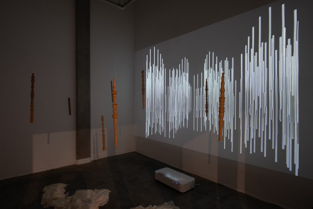 A dark gallery room displays many long, hanging forms, which are decorative wooden pole-like structures. Echoing these narrow forms, a projection of many thin, white forms is displayed on the wall. On the floor lay soft, white textile sculptures.