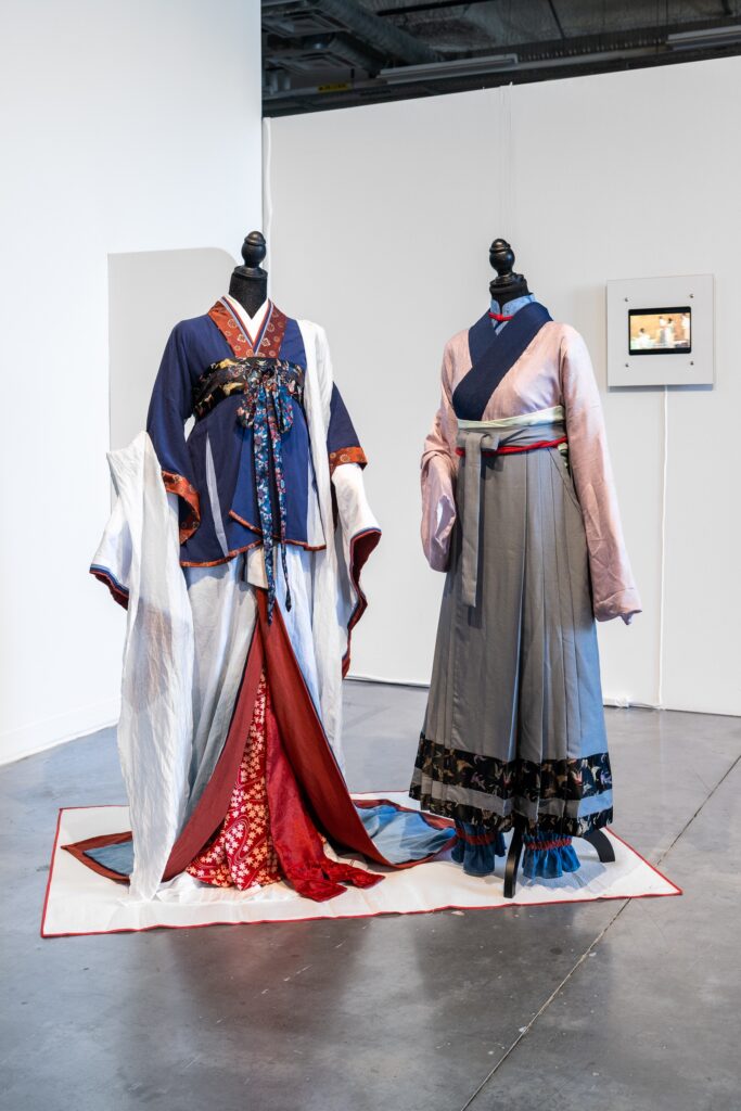 Two dressform mannequins display a set of traditional clothing, composed of a variety of colours and draped material. The mannequins stand on top of a rectangular textile sheet, matching the clothing. In the background, a small screen, featuring another work, is displayed on the gallery wall.