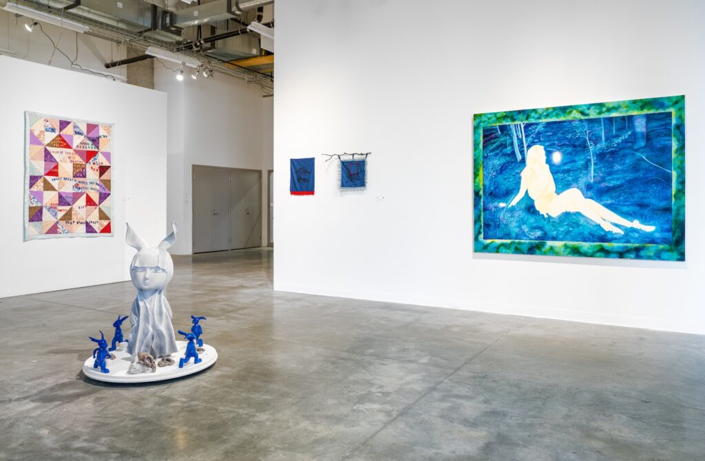 Multiple large scale, brightly coloured works are displayed on the wall and the floor of the gallery room. A quilted, geometric work hangs on its own wall. Made up of cool tones and a range of figures depicted in the works, the room is filled with a mystical, imaginative feeling. 