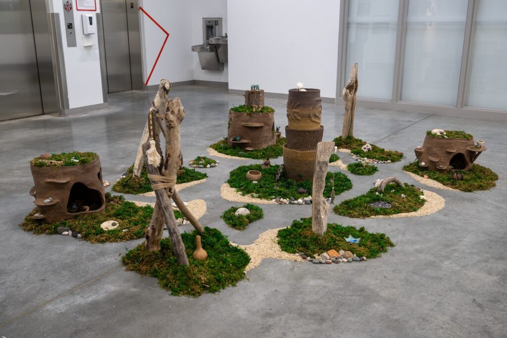 Shown up close, an archipelago-like installation covers the floor of the gallery room. The work is composed of mounds of organic material, including moss, sand, and stones, with a variety of objects sitting on top of each one. The objects, including wood, ceramic-like sculptures, and other ephemera, populate the earthy space.