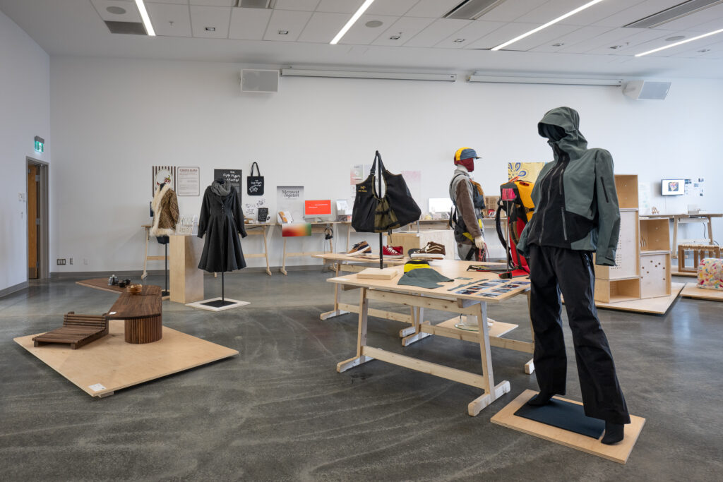 A bright gallery room shows a collection of design projects, including clothing, furniture, and posters. The works are displayed on mannequins, tables, and other display platforms. The works include athletic-type items, image-based media, shoes, innovative furniture, and more.