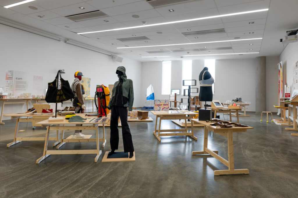 A bright gallery room shows a collection of design projects, including clothing, furniture, and posters. The works are displayed on mannequins, tables, and other display platforms. The works include athletic-type items, image-based media, shoes, innovative furniture, and more.