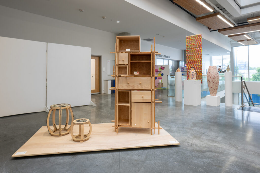 A bright gallery hall shows a collection of wood furniture projects, with ceramics and colourful hanging works in the background. The forms of the furniture are geometric and dynamic in height. The ceramics, standing on plinths, include vessels and figure-like forms.