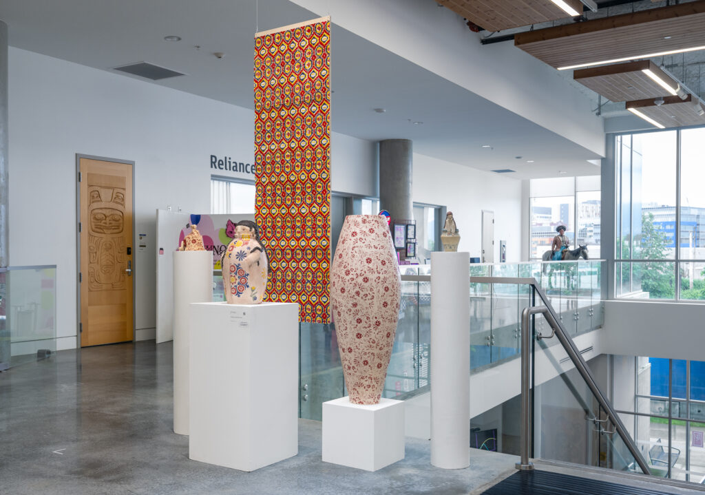 A bright gallery hall shows a series of ceramics and a colourful textile piece hanging in the background. The ceramics, standing on plinths, include vessels and figure-like forms. They range in size and are decorated with fine details and patterns.
