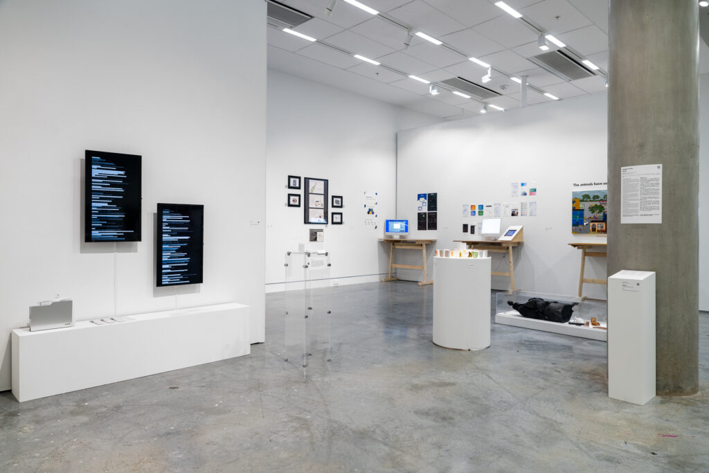 A gallery room shows a range of design projects, mostly digital and image-based. The projects are displayed on the wall, on plinths, and on tables, showing the depth of research and information of each work. The walls have screens, posters, and other images mounted on them.