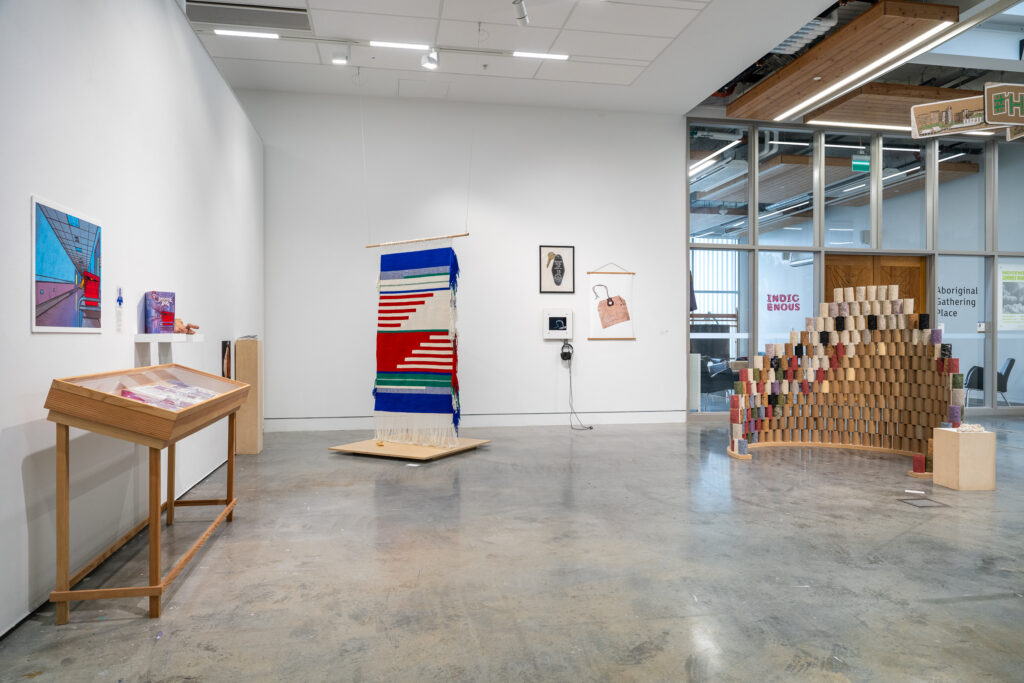 A gallery room shows a range of works, including a large hanging textile piece, a large sculptural installation, and multiple two-dimensional works on the walls. The room is filled with dynamic shapes and colours, with a variety of mediums displayed. 