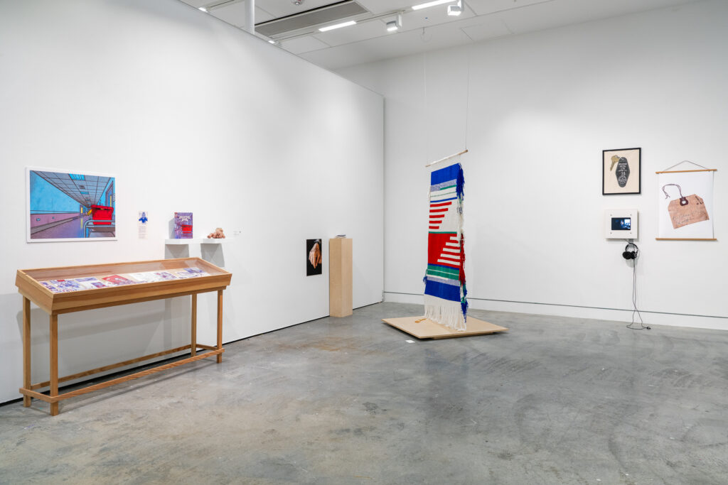 A gallery room shows a range of works, including a large hanging textile piece and multiple two-dimensional works on the walls. The room is filled with dynamic shapes and colours, with a variety of mediums displayed. 