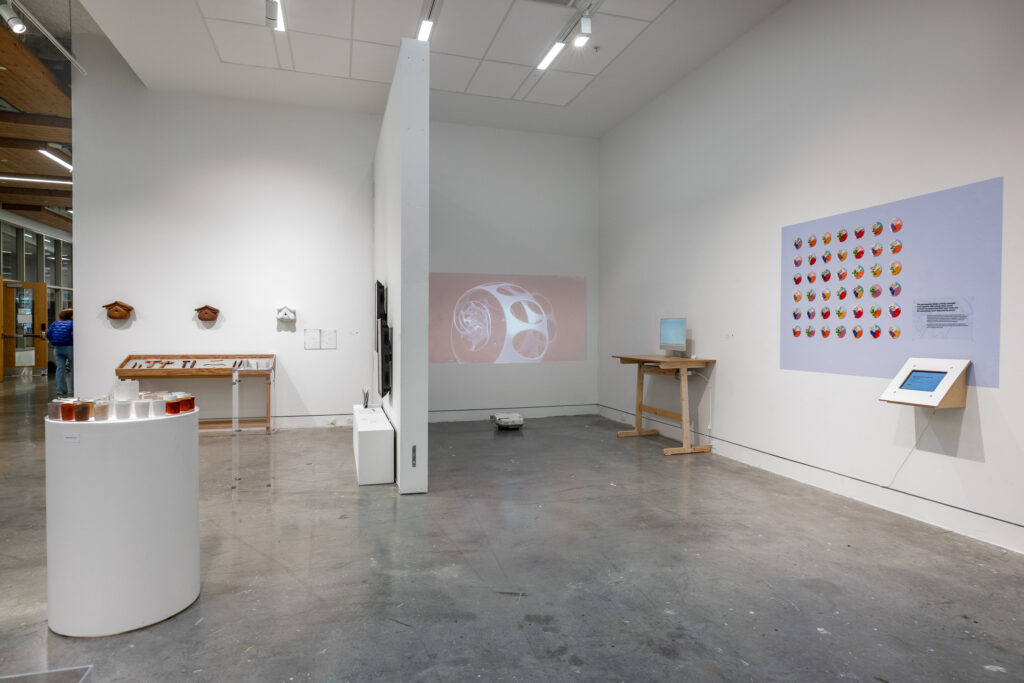 A gallery room, divided by a free-standing wall, displays a range of design projects. On one side of the dividing wall, the room is slightly darkened to highlight a projection on the wall, as well as projects displayed on a monitor and on a smaller mounted screen. A large colourful poster hangs on the same wall. On the other side of the dividing wall, there are design projects including wooden items, vessels, and more.