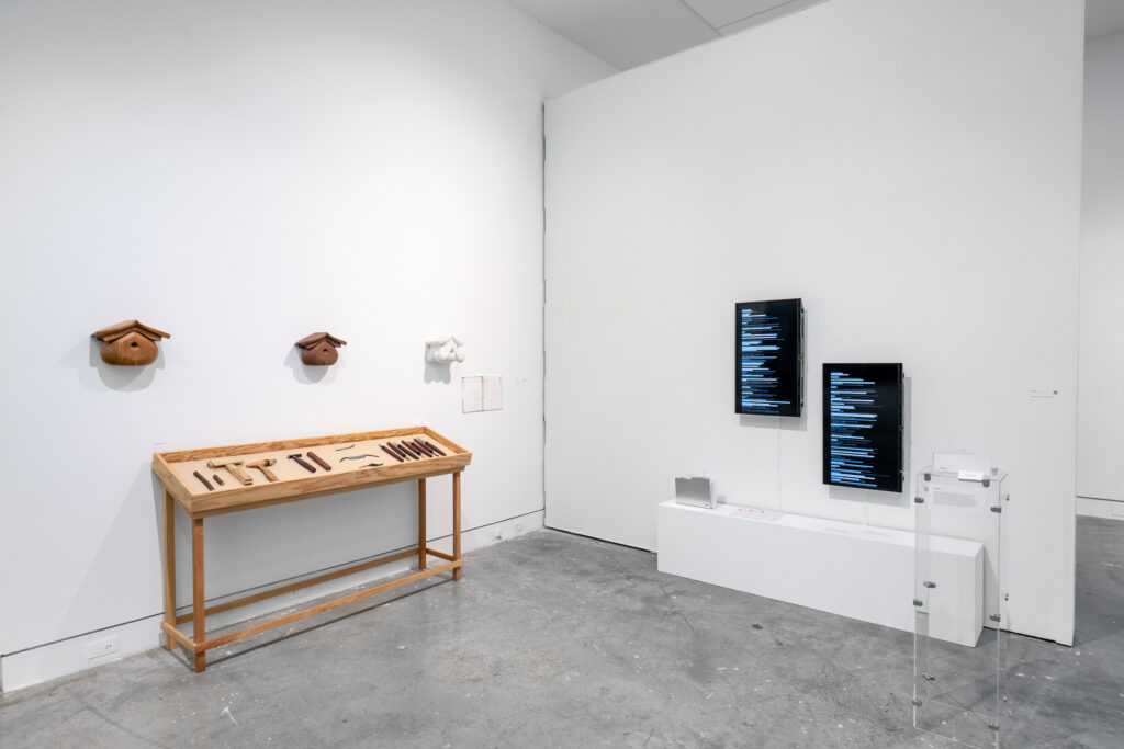 The gallery walls show a collection of wooden tools in a display case with wooden objects mounted above it. On the adjacent wall, there are two mounted screens showing lines of text. In the center of the room stands a transparent plinth with a small object on top.